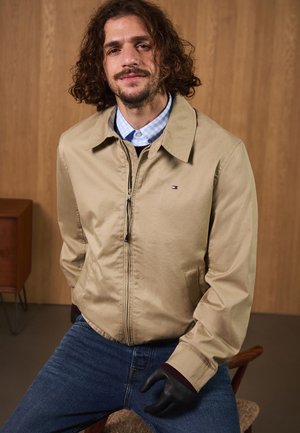 Beige bomber jacket with a zip closure, side pockets, and black knit cuffs. Worn over a blue checkered shirt, paired with blue jeans.
