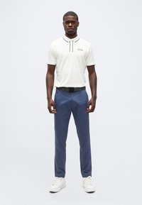 Άνδρας που στέκεται προς τα εμπρός, φορώντας λευκό πόλο πουκάμισο, μπλε παντελόνια, μαύρη ζώνη και λευκά αθλητικά παπούτσια σε απλό φόντο.