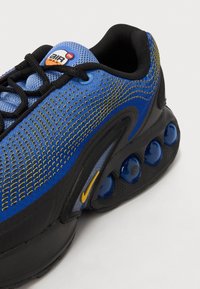 Chaussure de sport bleue et noire avec des accents jaunes, tige en mesh, coussins d'air visibles dans la semelle et lacets noirs sur fond neutre.