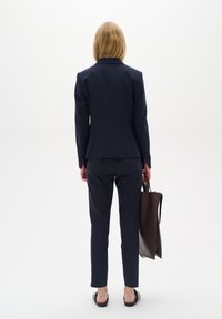 Personne aux cheveux blonds portant un costume bleu marine et tenant un sac fourre-tout marron foncé, debout dos à un fond blanc uni.