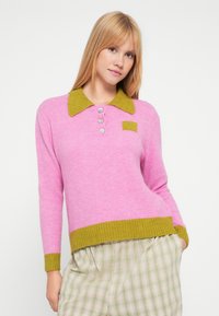 Maglione in lana rosa con collo a coste verde, patta con tre bottoni e una piccola toppa verde; abbinato a pantaloni beige a quadri.