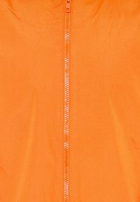 Giacca leggera arancione con zip completa, texture liscia e senza motivi. La zip è centrale con un cursore abbinato.