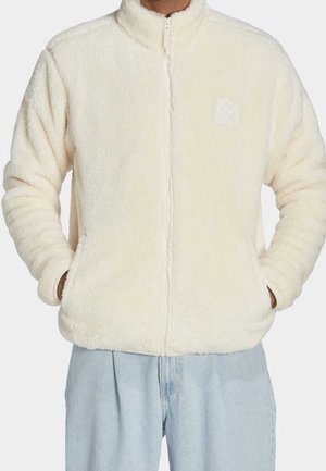 Person trägt eine cremefarbene, flauschige Jacke mit Reißverschluss und den Händen in den Taschen, kombiniert mit hellblauen Jeans.