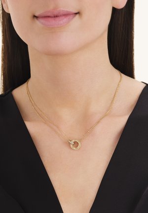 Close-up van een vrouw die een dubbele gouden ketting draagt met een cirkelvormige hanger met kleine ingelegde diamanten.