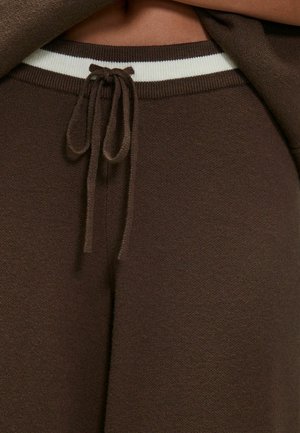 Braune Strick-Sweatpants mit einem weißen gestreiften Gummibund und einem Kordelzugverschluss. Die Textur ist weich und leicht dehnbar.