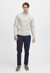 Light grey zip-up sweatshirt with ribbed cuffs and hem, logo on left chest, paired with dark navy trousers and grey shoes.