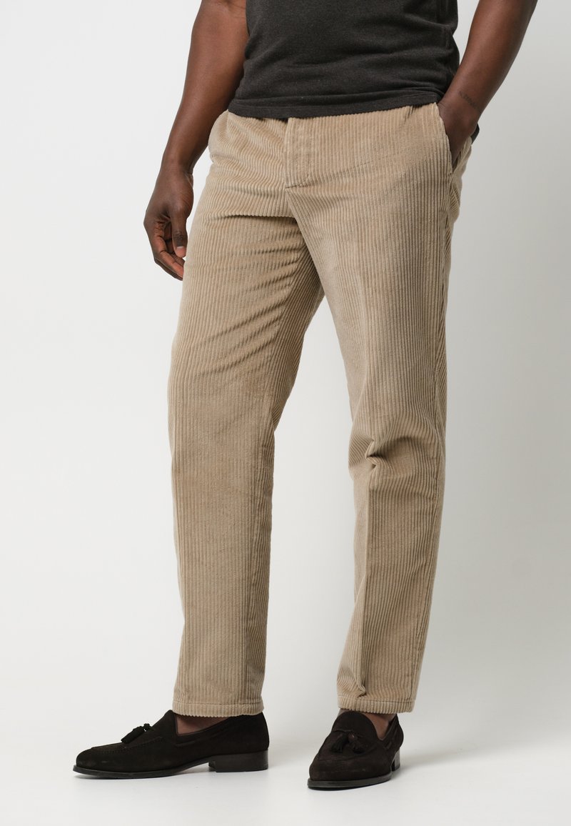 Beiga manchesterbyxor med vertikala ribbor, rak bendesign och sidofickor, ihop med mörka loafers för en komplett look.