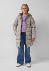 Djevojka nosi bež kaput s kapuljačom od jastučića, puderasto ljubičasti duksericu s printom grada, široke plave traperice i bijele tenisice, stoji uz jednostavnu pozadinu.