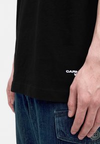 Gros plan de la main d'une personne reposant le long du corps, portant une chemise noire avec un texte blanc et un jean denim bleu foncé avec une poche boutonnée.