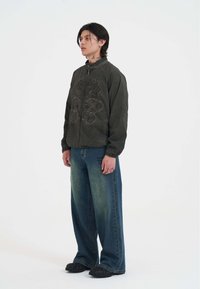 Grøn bomberjakke med broderet design, parret med løse blå denimjeans med lys vask og mørke sko.