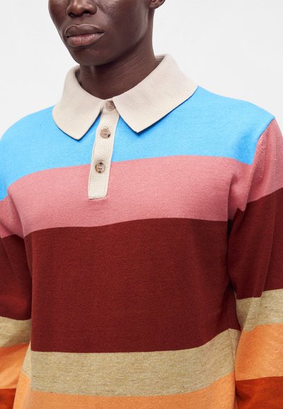 Homme portant un pull à col avec de larges rayures horizontales bleu, rose, rouge foncé, beige et orange.