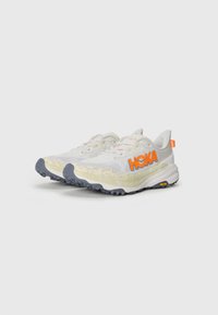 Scarpe sportive bianche con un design testurizzato, dotate di un logo arancione, suola grigia e marchio vibram; chiusura con lacci e collare imbottito.
