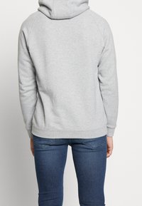 Šedá mikina s kapucí a volným střihem, s žebrovanými manžetami a lemem. Zadní pohled, minimální detaily, kombinováno s modrými džíny.