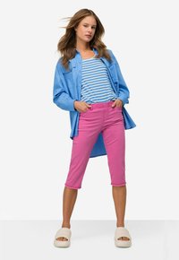 Camicia azzurro chiaro con bottoni sopra una canotta a righe blu e bianche, abbinata a pantaloni cropped rosa e sandali beige.