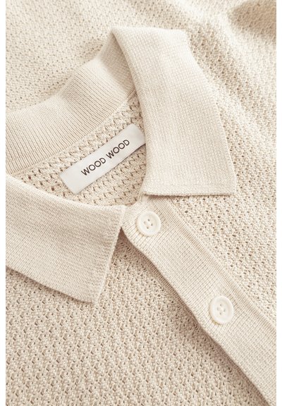 Polo en tricot beige avec un design texturé, un col classique et deux boutons blancs, avec une étiquette portant la mention "WOOD WOOD."