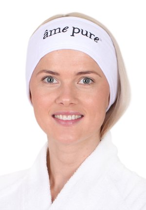Hvit hodebånd med svart tekst "âme pure" bæres av en person med lyseblondt hår, smilende, i en hvit robe mot en ensfarget bakgrunn.