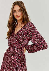 Čierne šaty s dlhým rukávom a ružovým kvetinovým vzorom, s viazaním v páse a výstrihom do V. Ľahká, textúrovaná tkanina.