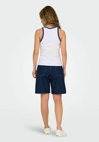 Top bianco senza maniche con bordi blu navy, abbinato a pantaloni corti in denim scuro. Calzature casual completano l'outfit, mettendo in mostra un fit e un design rilassati.