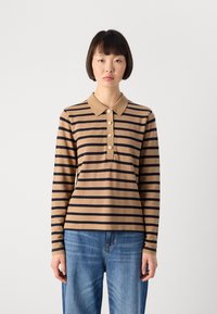 Giovane donna con capelli corti e scuri che indossa una polo a maniche lunghe a righe marroni e nere e jeans blu, davanti a uno sfondo semplice.