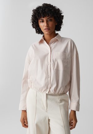 Frau mit lockigem Haar, die ein hellrosa gestreiftes Hemd mit Knöpfen und hoch taillierte cremefarbene Hose trägt, steht vor einem schlichten grauen Hintergrund.