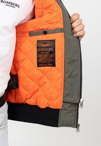 Blouson aviateur matelassé avec intérieur orange, extérieur gris, poignets côtelés et une étiquette noire avec la marque et les détails. Fermeture éclair incluse.