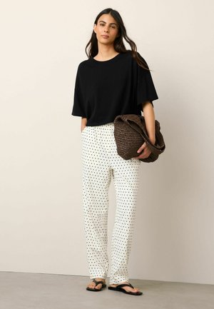 Woman standing with hands in pockets, wearing black oversized t-shirt, white polka dot pants, black sandals, and holding a brown woven bag.