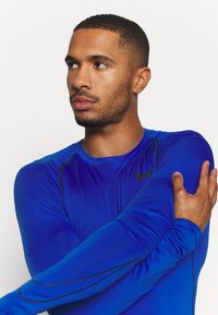 Langärmliges Sportshirt aus lebhaft blauem Stoff, mit schwarzen Akzenten und taillierter Passform. Glatte Textur mit leichtem Glanz. Nike-Logo sichtbar.