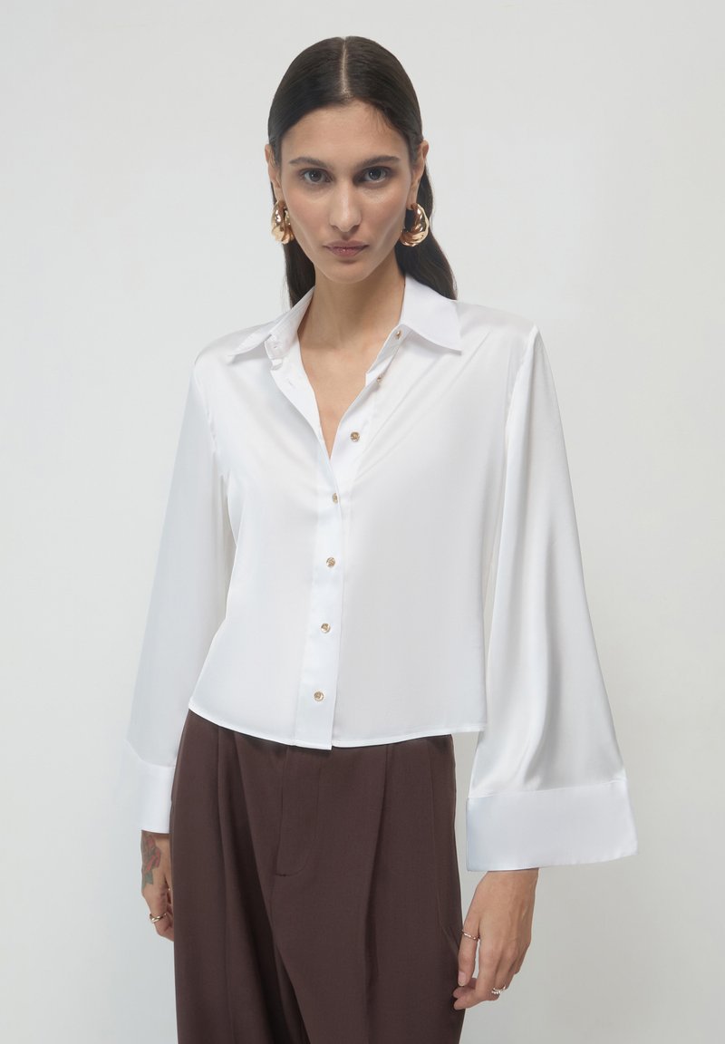 Donna con lunghi capelli scuri che indossa una camicetta bianca in satin con bottoni e ampi polsini, abbinata a pantaloni marroni a vita alta, su uno sfondo semplice.