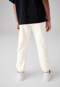 Cremefarvede sweatpants med elastiske manchetter, lavet af blødt stof, parret med hvide sneakers, set fra ryggen.