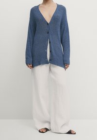 Blått stickat cardigan med djup V-ringning, knappstängning fram, ribbade ärmslut och nederkant. Kombineras med vita vida byxor och svarta sandaler.