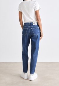 Džínsy s vysokým pasem, modré, se rovnými nohavicemi, denimová látka, dvě zadní kapsy, opasek s logem, nošeny s bílými platformovými teniskami.