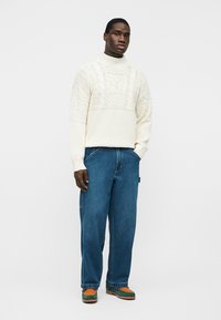 Mann steht mit einer Hand in der Tasche, trägt einen weißen, strukturierten Strickpullover, lockere blaue Jeans und mehrfarbige grüne und orangefarbene Schuhe.