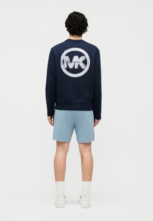 Person med krøllet hår iført en marineblå sweatshirt med en forvrænget hvid cirkulær logo, lyseblå shorts, hvide sokker og hvide sneakers, set bagfra.