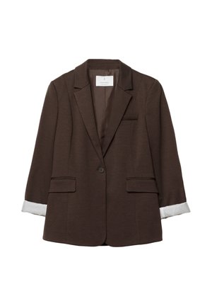 Femme portant un blazer marron foncé ajusté avec des manches blanches retroussées, associé à un pantalon clair, vue de profil et de dos.
