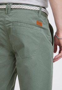 Pantalones de algodón verde claro con dos bolsillos traseros, una etiqueta de cuero marrón y un cinturón trenzado blanco visible en la cintura.