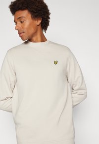 Világos bézs crewneck pulóver hosszú ujjal, bal mellen hímzett sárga pillangó logóval. Sima textúra.