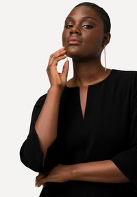 Schwarzes Kleid mit V-Ausschnitt und Dreiviertelärmeln, das eine glatte Textur aufweist. Das Model trägt lange, hängende Ohrringe und einen neutralen Lippenstift.