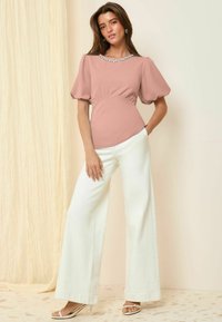 Blusa rosa cipria con maniche a sbuffo e accentatura di gioiello nel colletto, abbinata a pantaloni bianchi a gamba larga e sandali leggeri. Texture del tessuto liscia.