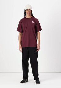 Bordeaux bomulds-t-shirt med et pink hjerte-logo på venstre bryst, parret med løstsiddende sorte bukser og en lys beige kasket.