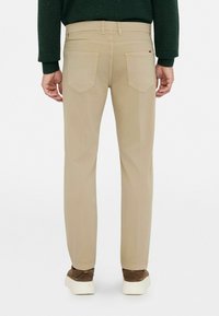 Chinos beige con un corte ajustado, que cuentan con dos bolsillos traseros, costuras visibles y un sutil acento de logo cerca de la cintura. Combinados con zapatillas marrones.