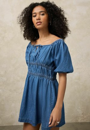 Frau mit lockigem Haar, die ein blaues Off-Shoulder-Jeanskleid mit Puffärmeln trägt und vor einem neutralen Hintergrund steht.