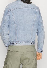 Ljusblå denimjacka med en figurnära design, synlig sömnad och två bruna tryckknappar i midjan, bärs över olivgröna cargobyxor.