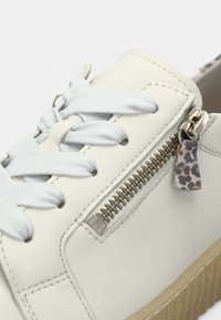 Valge nahast toss siniste paelte, tõmblukukaunistuse ja väikese leopardimustrilise detailiga. Tekstureeritud tan-nahkne kummitald. Lähedalt vaadatuna.