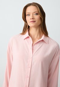 Vrouw met lichtbruin haar die een lichtroze knoopshirt draagt tegen een effen lichtgrijze achtergrond.