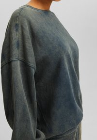 Sudadera de gran tamaño en un color azul-verde desvaído, hecha de algodón suave, con cuello redondo, hombros caídos y costuras prominentes.