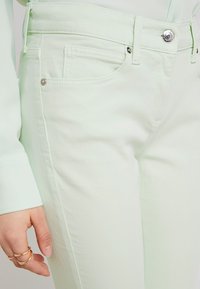 Acercamiento a unos pantalones verde claro con botón de metal y remaches, bolsillo delantero, y una mano que lleva dos anillos de oro descansando al lado de la pierna.