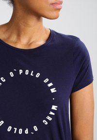 Žena oblečená v námořnicky modrém tričku s bílým kruhovým textem "Marc O'Polo DNM" na přední straně, zachycena od ramen po střed hrudníku.