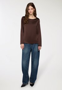 Top lungo a maniche lunghe in satin marrone abbinato a jeans in denim blu a gamba larga e scarpe nere a punta, su uno sfondo neutro.