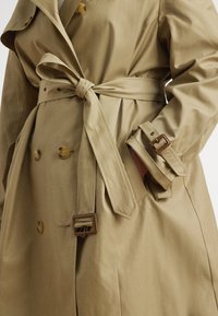 Bézs színű ballonkabát övvel a deréknál, kétsoros gombolással, nagy gombokkal és mandzsettabeállítással, sima, könnyű anyagból.