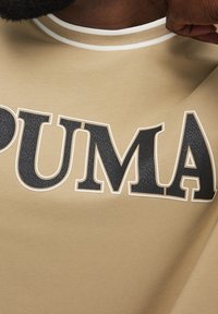 Primer plano de una persona estirando la tela beige de una camiseta con grandes letras negras texturizadas "PUMA" en el pecho.